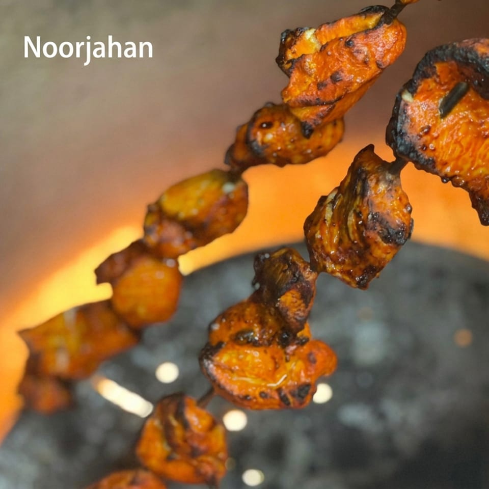 Charred orange chicken skewers suspended over glowing charcoal, with Noorjahan text visible in the upper left.