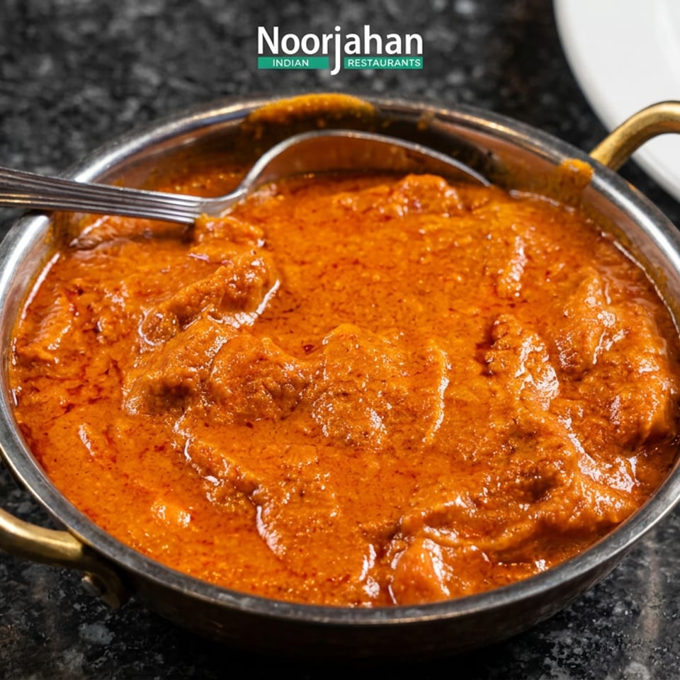 Creamy orange Indian curry with tender pieces in a brass-handled serving bowl and spoon on a dark countertop.